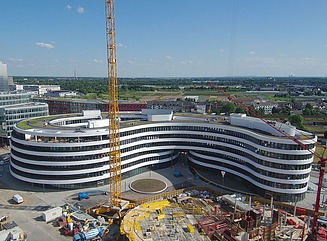 Helles Gebäude mit geschwungener Fassade in 6 Geschossen, links Promenade mit Menschen, die an Tischen sitzen oder flanieren