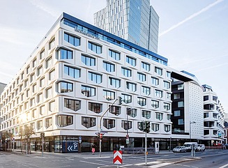 Eckgebäude mit strukturierter Außenfassade, im erdgeschoss Ladengeschäfte, Baustellenbegrenzung im Vordergrund, Hochhaus Skyline im Hintergrund