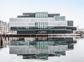 Futuristisk bygning lavet af flere grønlige glasblokke med et gennemgående hvidt gulv ved en vandmasse 