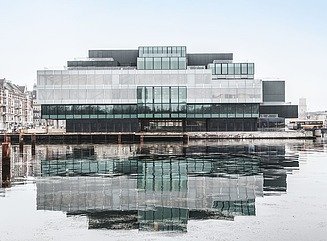Futuristisk bygning lavet af flere grønlige glasblokke med et gennemgående hvidt gulv ved en vandmasse 