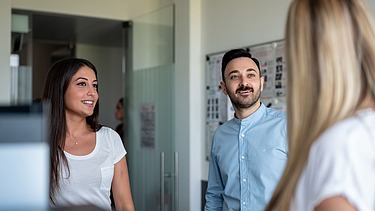 A blonde woman from behind, a man and a woman from the front talking to each other in front of a metaplan wall