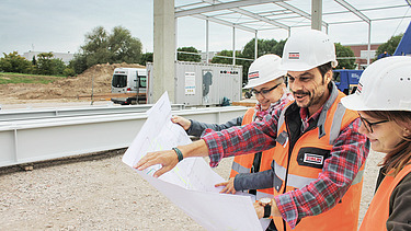 View of a plan in front of which three people with construction helmets are standing and talking about it
