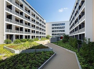 Bred plads med et par mennesker i forgrunden, i baggrunden en studenterbolig med en struktureret hvid facade foran en beigefarvet bygning