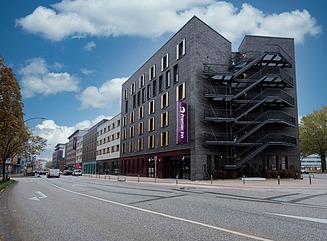 Eckgebäude an einem Platz mit grauer Fassade, fünf Außentreppen und schmalen Fens
