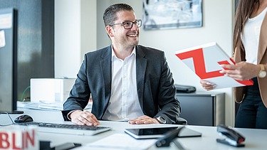 Man in a suit at a desk talks to a person holding a folder with the Züblin logo on it