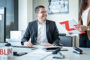 Man in a suit at a desk talks to a person holding a folder with the Züblin logo on it