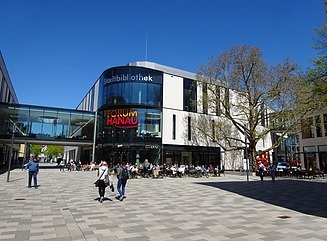 Plads med forbipasserende foran to bygninger, der er forbundet med en søjlegang. Bygningen til venstre har store vinduer, bygningen til højre har gule områder ved siden af vinduerne.