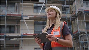 Frau mit Helm und orangefarbener Weste steht vor einem Gerüst und hält ein Pad in der Hand