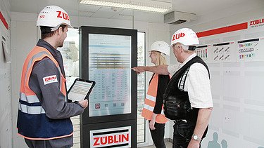 Three people in orange vests discuss a process diagram in front of a screen