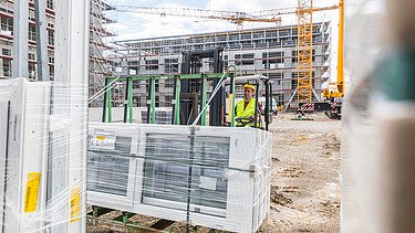 Mann in gelber Weste auf einem Gabelstapler mit Glasfenstern. Baustelle mit gelbem Kran im Hintergrund