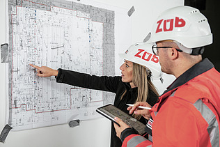 View of a plan in front of which two people with construction helmets are standing and talking about it