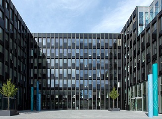 Bürokomplex mit dunkler Fassade in U-Form um einen gepflasterten Platz, rechts zwei türkisfarbene Stelen