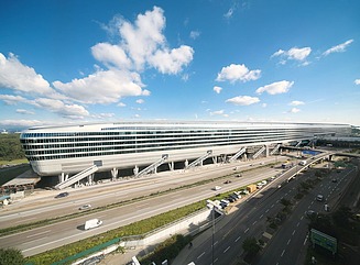 Four-lane road with red tail lights visible through long exposure, green strip on the right, behind it an elongated building complex that widens towards the top, with a rounded façade 