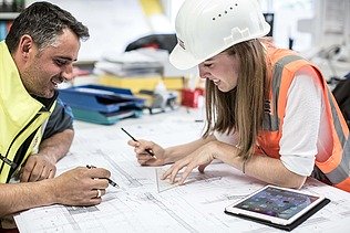 Ein Mann in gelber Weste und eine Frau mit Helm und orangefarbener Weste arbeiten gemeinsam an einem Plan, auf dem Tisch ein Pad