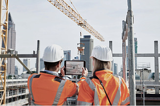 Construction site contract models Two people wearing helmets and orange vests inspect a plan on a pad in front of a construction site with a crane