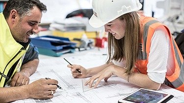 Ein Mann in gelber Weste und eine Frau mit Helm und orangefarbener Weste arbeiten gemeinsam an einem Plan, auf dem Tisch ein Pad