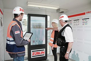 Three people in orange vests discuss a process diagram in front of a screen