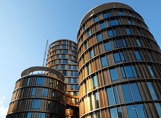 Vier versetzte runde Türme mit unterschiedlich großen erleuchteten Fenster, davor Bäume und einige Passanten