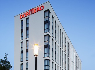Weißes Hotelgebäude mit dem Schriftzug Dormero in rot, auf der rechten Seite gelbe Sonnenschirme