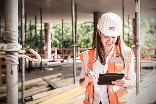 Junge Frau mit Helm und langen Haaren schaut auf einer Baustelle auf ein Pad