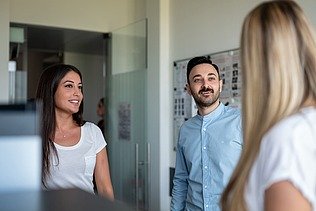 A blonde woman from behind, a man and a woman from the front talking to each other in front of a metaplan wall