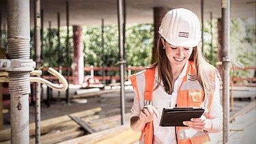 Junge Frau mit Helm und langen Haaren schaut auf einer Baustelle auf ein Pad