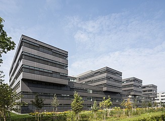 Dreigschossiger Sockelbau mit mehreren dreigschossigen Aufbauten mit durchlaufenden Fensterfronten, Grünfläche mit Bäumen im Vordergrund