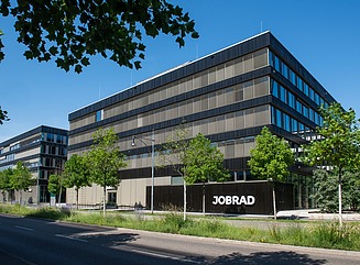 Corner building with horizontal window design, black cladding, construction fence in front of the building with the inscription Milestone