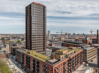 Hochhaus umgeben von einem u-förmigen Wohnblock mit begrünten Dachflächen, dahinter Stadtpanorama mit Kran