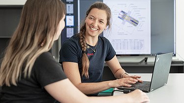 Lächelnde Frau am Laptop vor einem großen Screen mit Bauplänen im Hintergrund