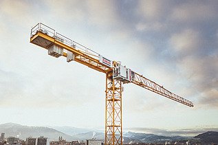 Kran vor einem Wolkenhimmel. Am unteren Bildrand Teile einer Stadt mit Hochhäusern vor Bergpanorama