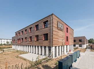 Mehrere Wohngebäude mit Holzverkleidung und ziegelroten Akzenten an den zwei Geschossen oberhalb des Erdgeschosses.