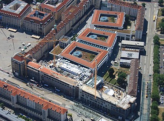 Viergeschossiges Eckhaus im Gründerzeitstil mit Arkadengang auf einem weitläufigen Platz mit Autos und Passanten im Vordergrund