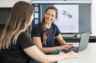 Lächelnde Frau am Laptop vor einem großen Screen mit Bauplänen im Hintergrund