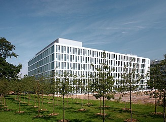 Hvidt kontorkompleks med mange smalle vinduer, græsplæne med flere rækker træer i forgrunden