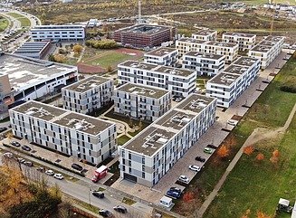 Fugleperspektiv af flere boligblokke med en sportsplads og et L-formet kompleks omkring en plads, omgivet af parker