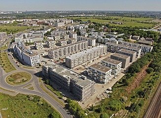 Vogelperspektive auf Mehrfamilienhäuser, ein heller Riegel mit schmalen Fenstern an einer Straße, dahinter mehrere Blocks mit Grünflächen dazwischen.
