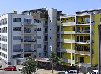 Straßenansicht mit weißem Eckhaus mit vorgebauten Balkonen, auf der rechten Seite gelb gestrichener Bauflügel mit umlaufenden Balkonen