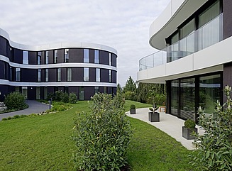 Leicht ansteigender Weg mit Rasenflächen an beiden Seiten zu einem Gebäude mit horizontaler weißer Struktur, schmalen Fenstern in schwarzer geschwungener Fassade