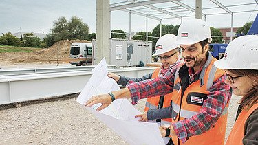 Udsigt over en plan, som tre personer med byggehjelme står foran og taler om