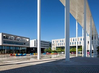 Lys blokbygning med fire etager og butiksvinduer i stueetagen, vinduerne på de øverste etager med hvide paneler. Et smalt tag på høje søjler fører over pladsen