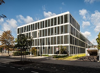 Bürogebäude mit großen Fenstern Verkehrsschilder und parkendes Auto, Bäume im Vordergrund