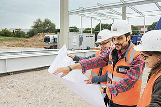 Blick auf einen Plan vor dem drei Personen mit Bauhelmen befinden und darüber sprechen