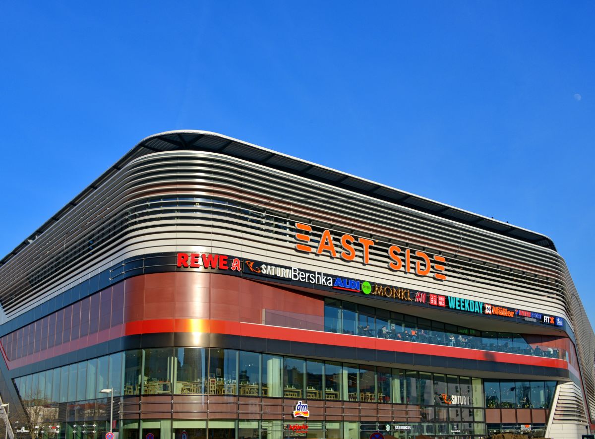 Gerundete Fassade eines Einkaufscenters mit dem Schriftzug East Side, hohen Fensterfronten im Erdgeschoss und roten Elementen darüber