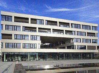 Weißes Gebäude mit 5 Geschossen, Ladenzeile im Erdgeschoss und ein Durchbruch über zwei Etagen in in der Mitte, vor dem Gebäude in Brunnen.