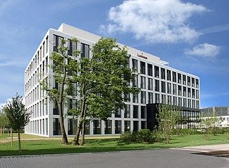 Lawn with trees and shrubs in the foreground, behind it a five-story white office building complex with large windows