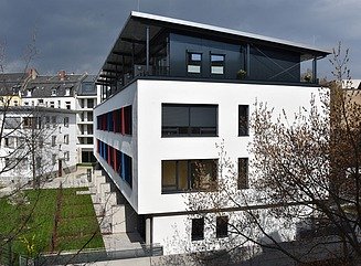 Weißes Wohngebäude mit vielen Fenster, teiweise rot undblau akzentuiert, Dachgeschoss mit schrägem Dach auf Säulen