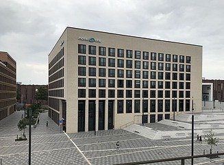 Grauer Hotelblock mit quadratischen Fenstern in den oberen Stockwerken und Motel One-Schriftzug, davor ein Platz mit mehreren Stufen