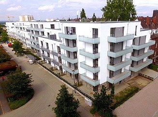 Weißes Wohngebäude mit 5 Geschossen und grauen Balkonen, im Vordergrund einige Bäume