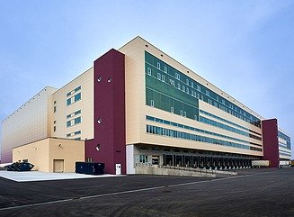 Light-colored building with dark red building elements, delivery ramps on the first floor, offices above with windows arranged in rows